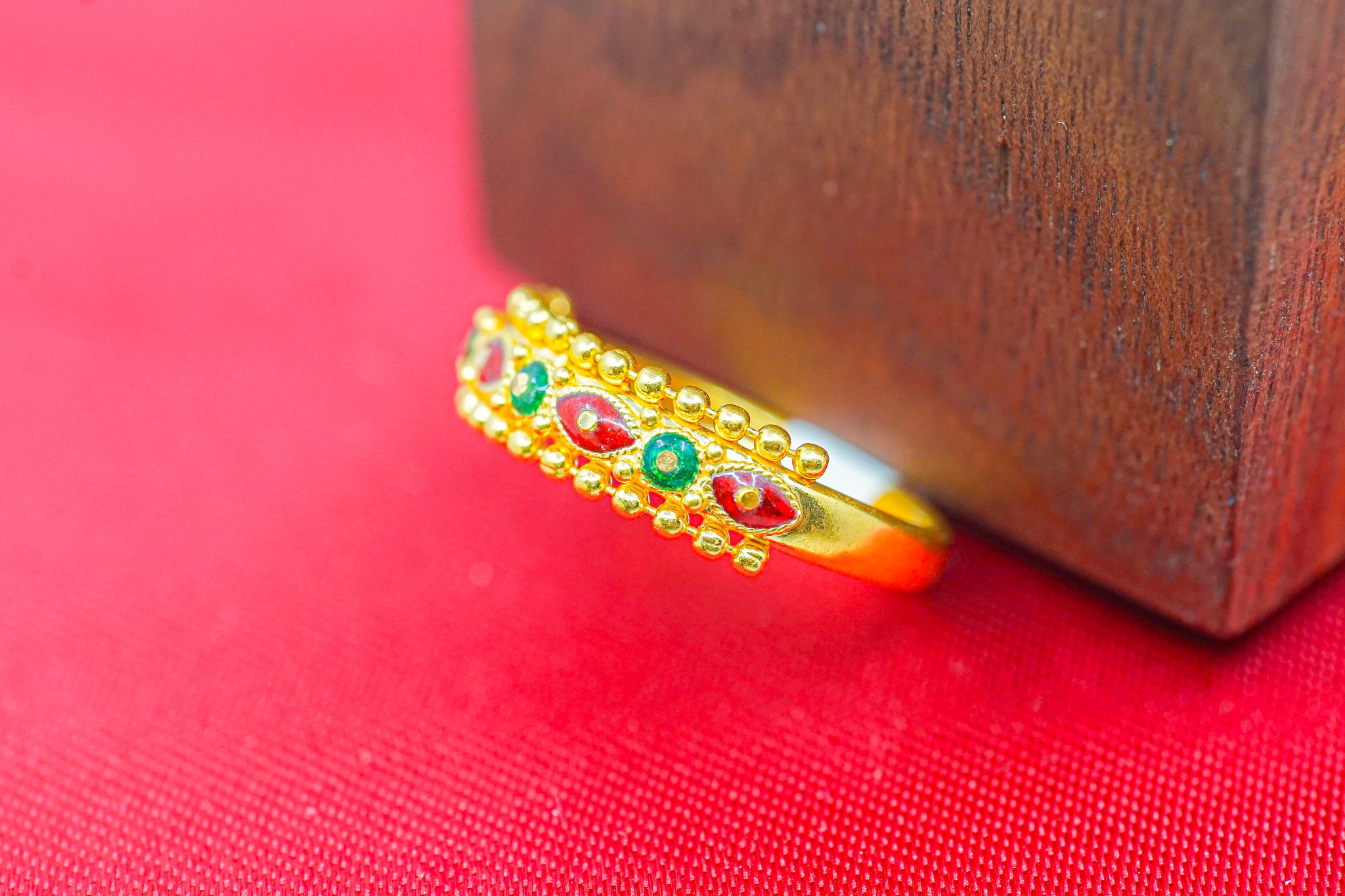 Close-up of a 22k gold beaded band ring with red and green accents on a red surface.