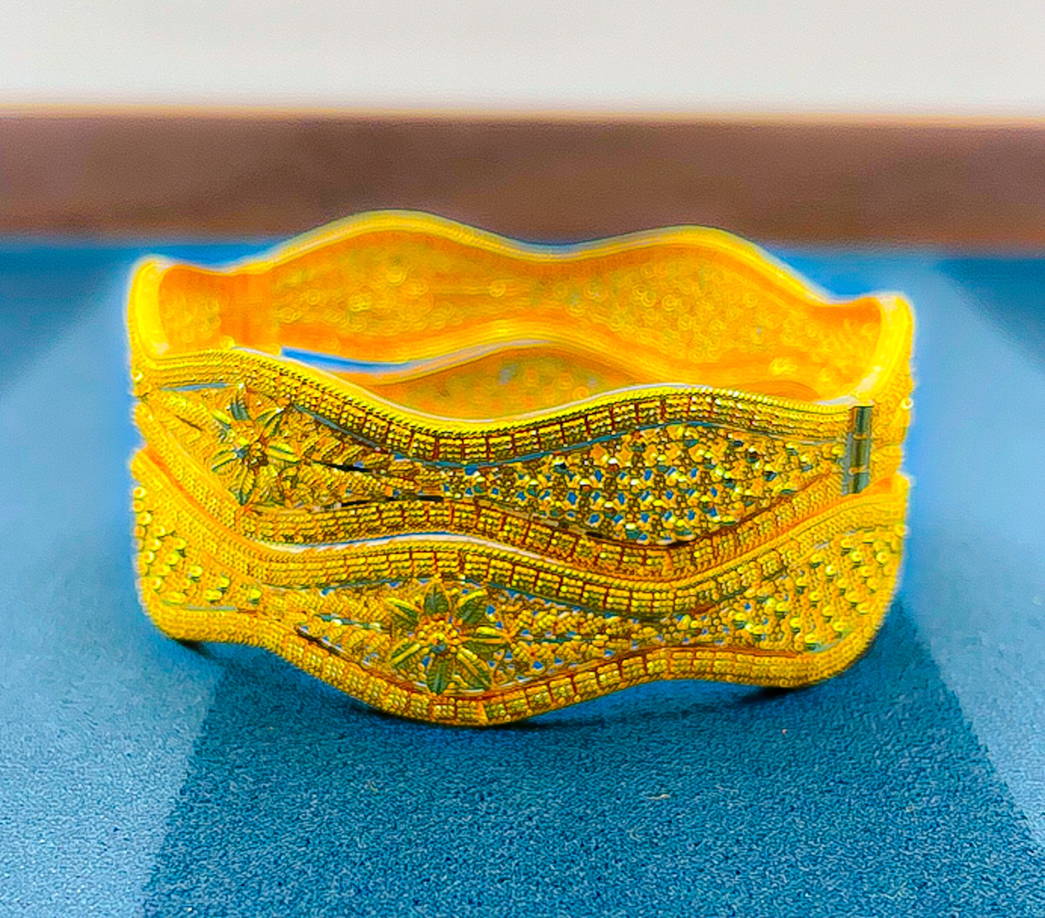 Wide 22k yellow gold bangle with intricate traditional patterned detailing photographed on blue surface
