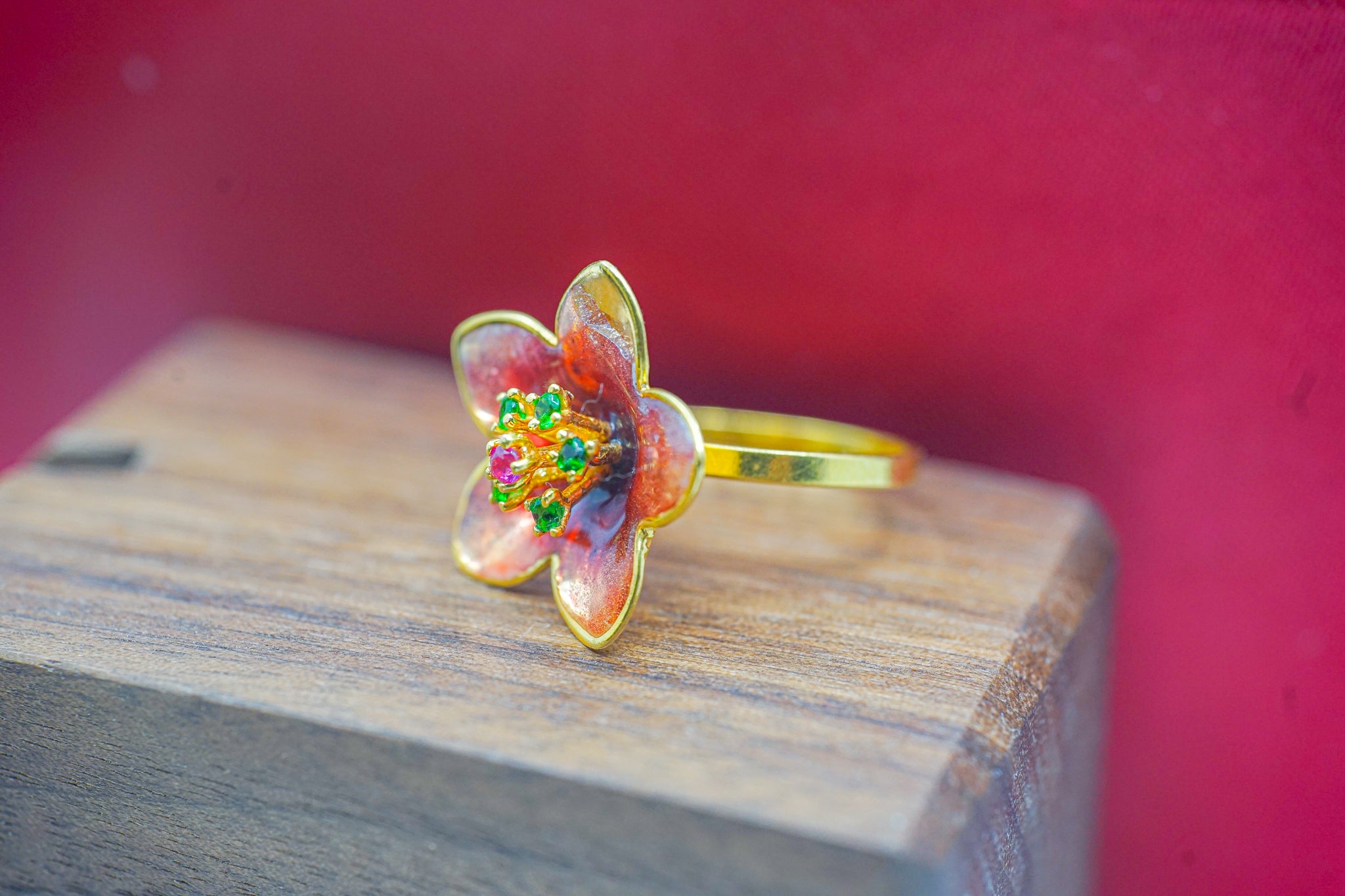 22k gold flower ring with red and orange translucent petals and green and pink accents on a wooden surface