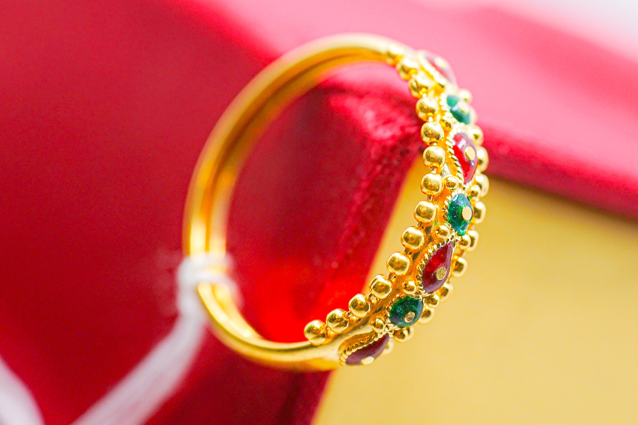 22k yellow gold ring with small red and green stones, photographed on a red and yellow background.