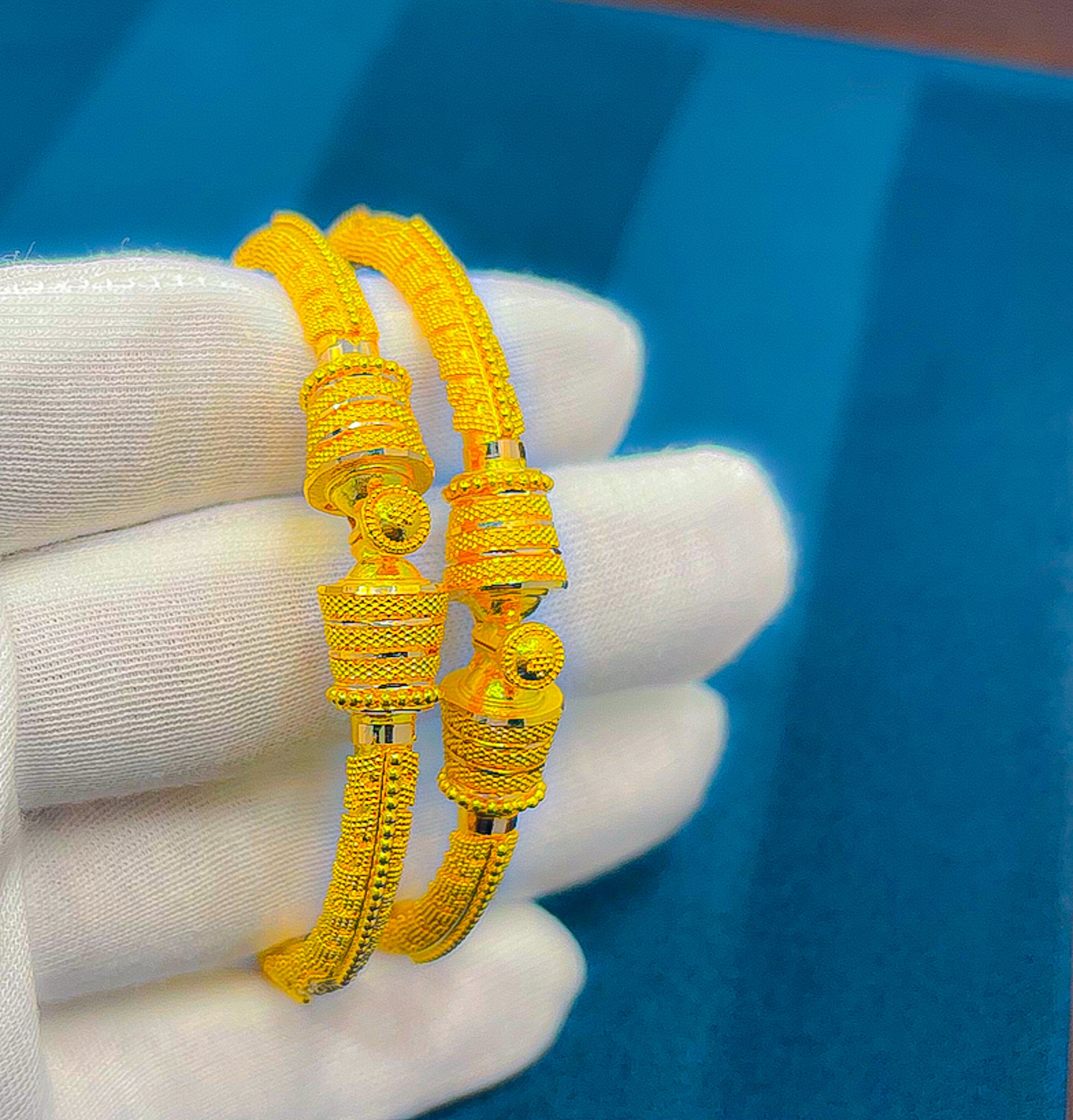 Close-up of a pair of 22k yellow gold bangles held in a white glove showing the textured design and center detail.