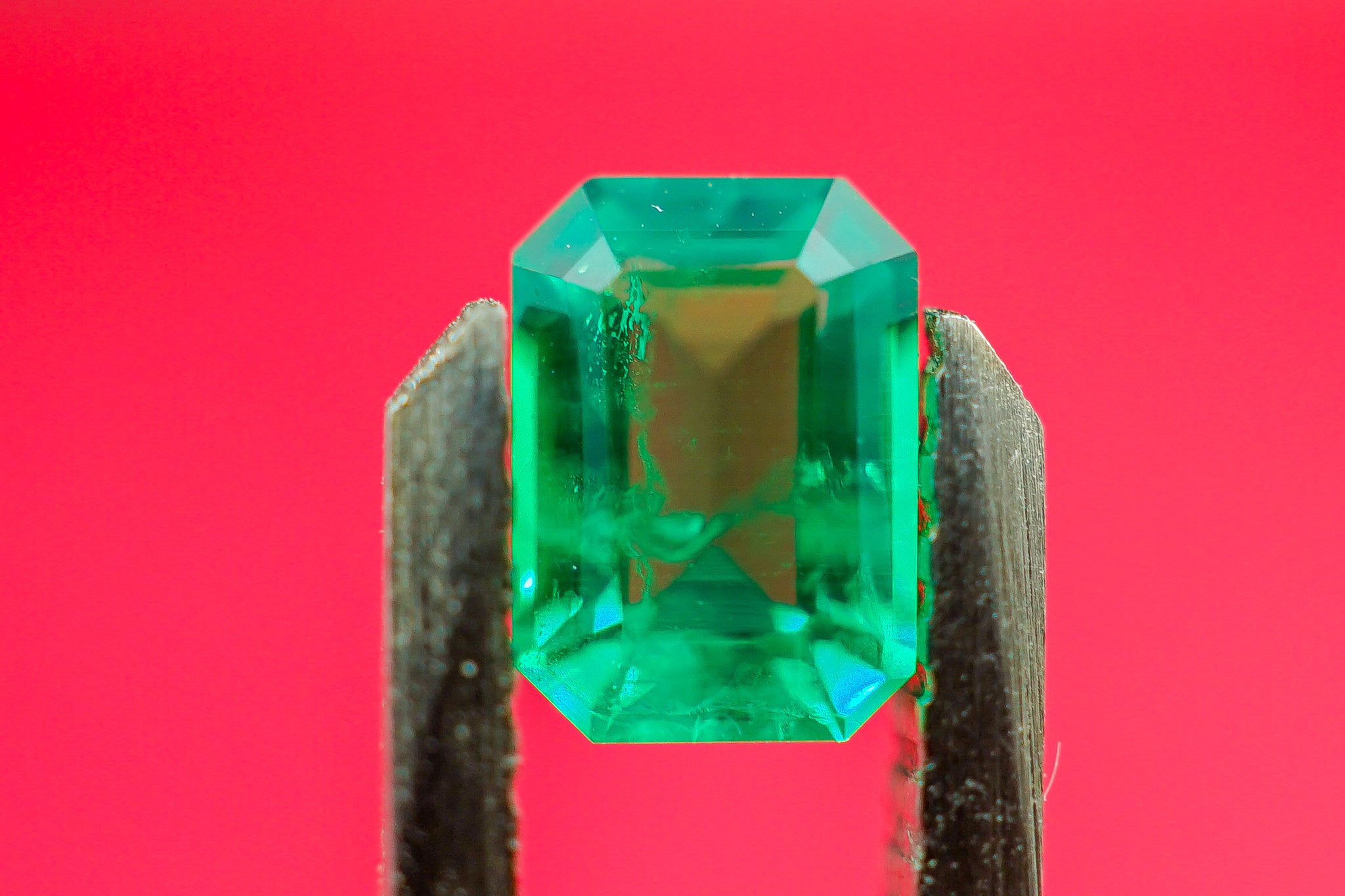 Green emerald gemstone 0.17 g held in tweezers showing emerald cut shape against red background