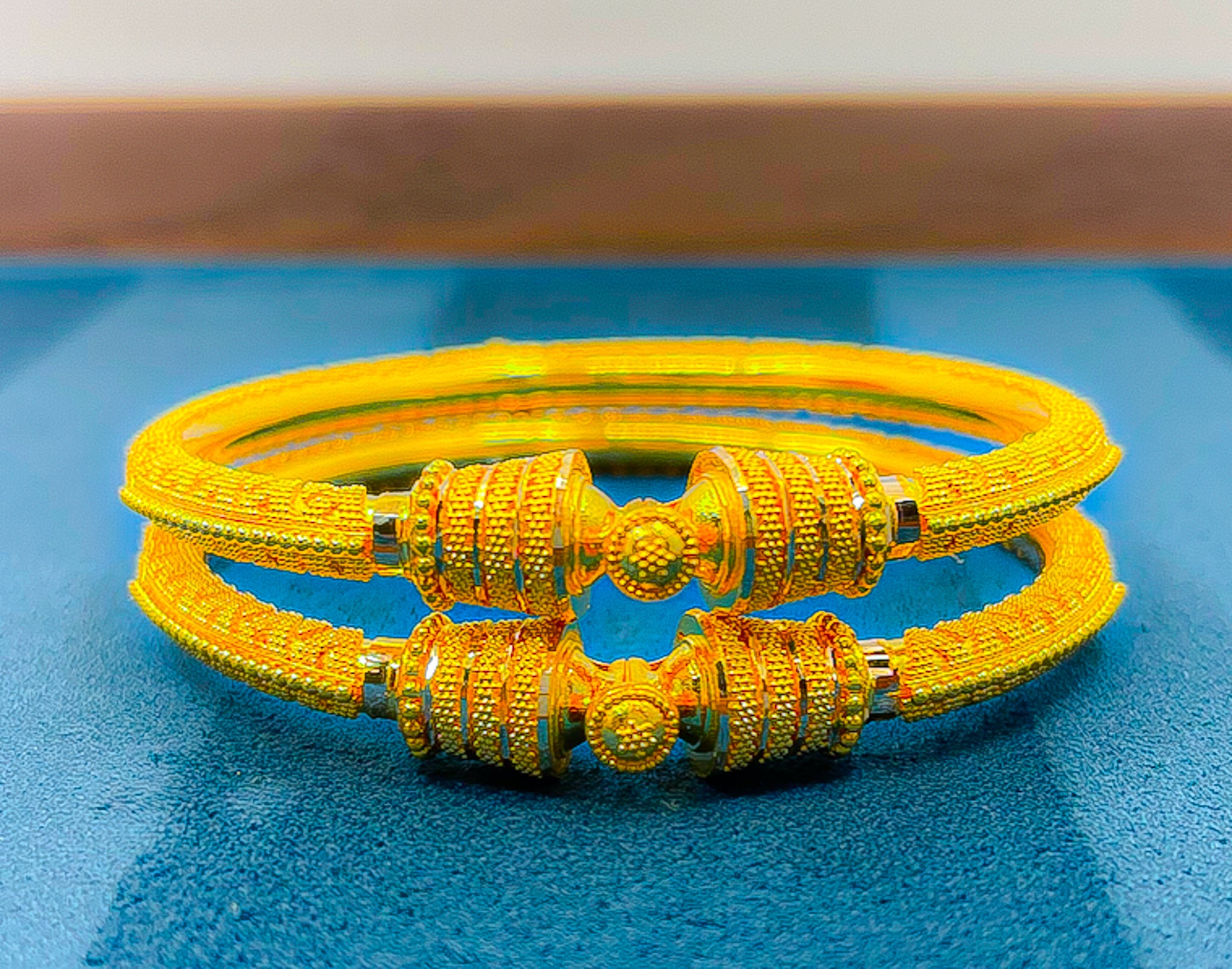 Pair of 22k yellow gold bangles photographed from an angle on a blue background showing the detailed texture.