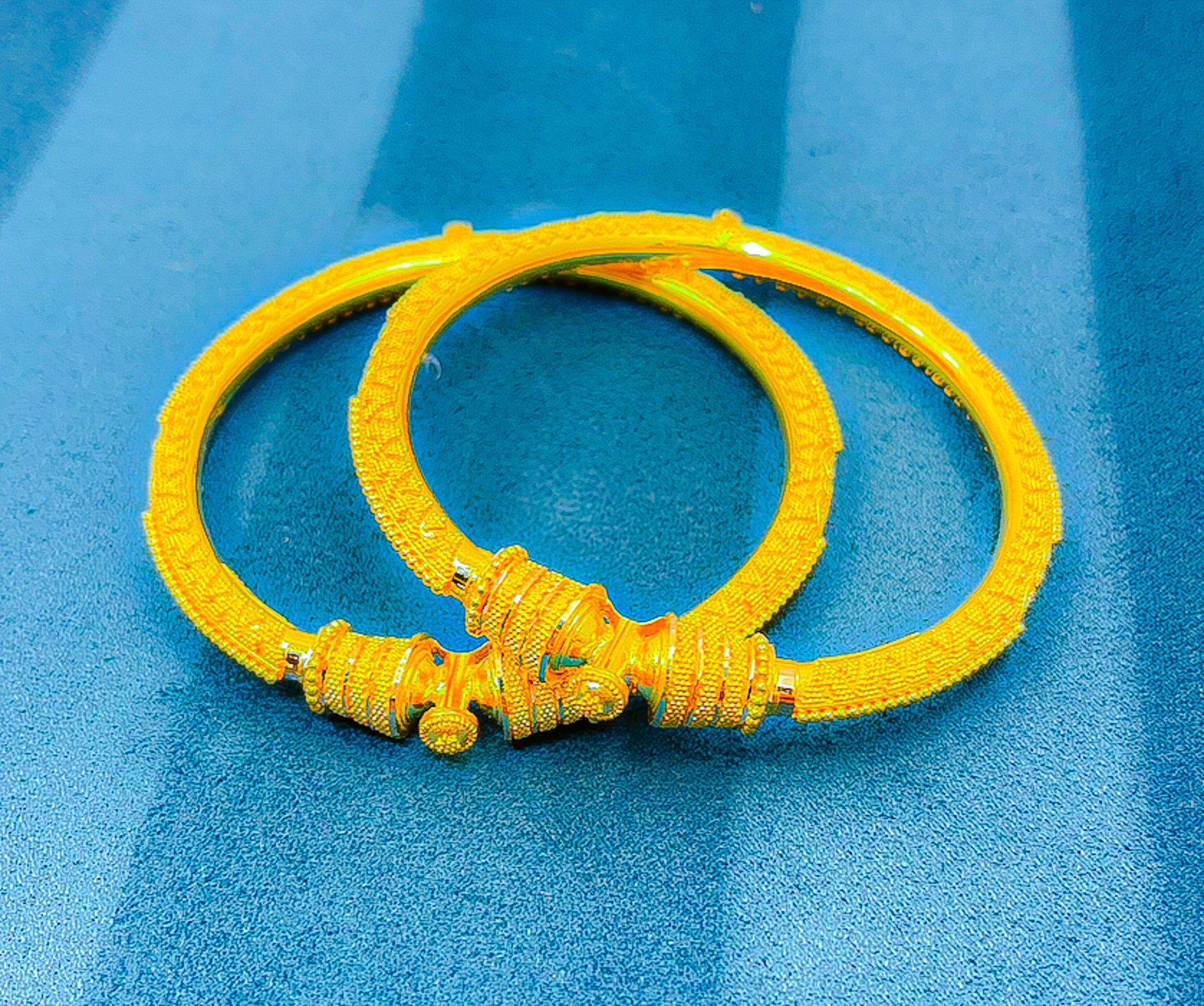 Pair of 22k yellow gold bangles photographed from an angle on a blue background showing the detailed texture.
