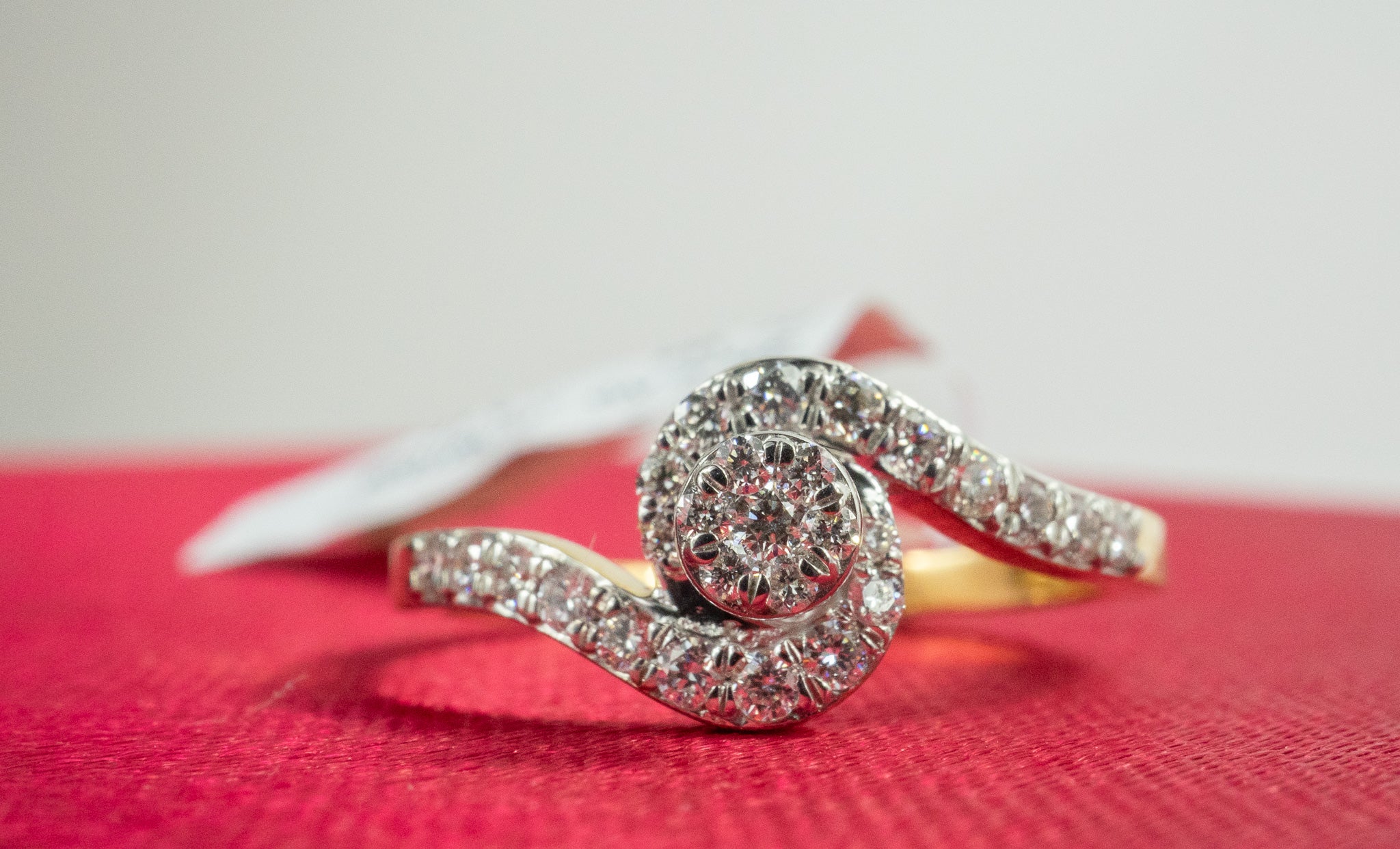 Glimmer Dot Ring in 18K gold with 0.23 carat diamonds in a swirl bypass design, photographed on a red background.